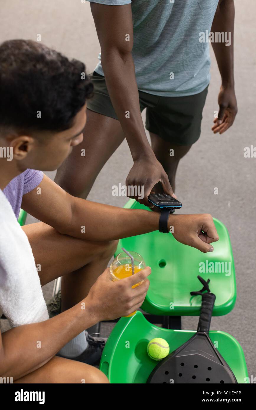 Divers amis masculins se reposant et regardant smartwatch sur le terrain avec raquette de paddle, boisson sportive Banque D'Images