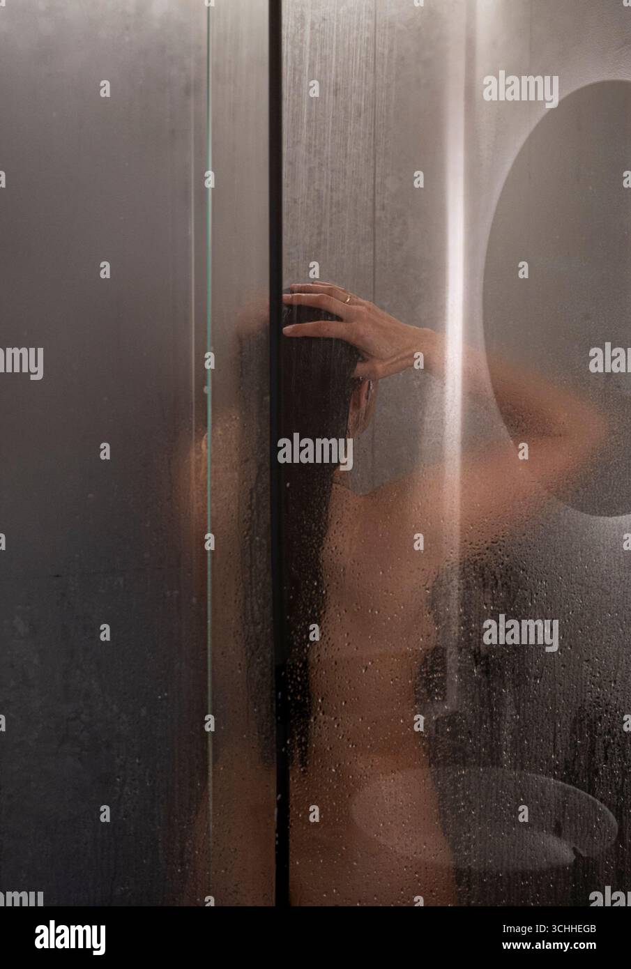 Une femme se tient debout sous une douche avec les cheveux mouillés. La douche est brumeuse et la femme se regarde dans le miroir Banque D'Images