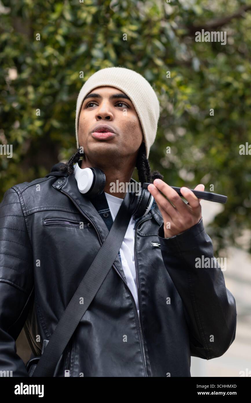 Homme afro-américain dans la vingtaine parlant dans un smartphone à l'extérieur en bonnet, écouteurs Banque D'Images