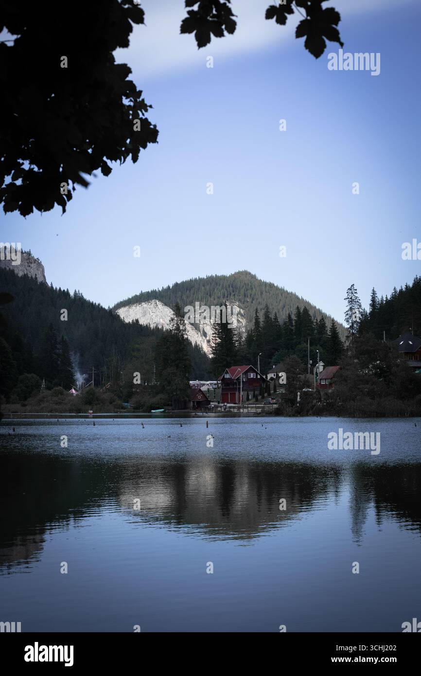 Un paisible village de montagne avec des maisons au toit rouge se trouve au bord d'un lac, son reflet reflet dans l'eau calme. De denses forêts de pins couvrent les environs Banque D'Images