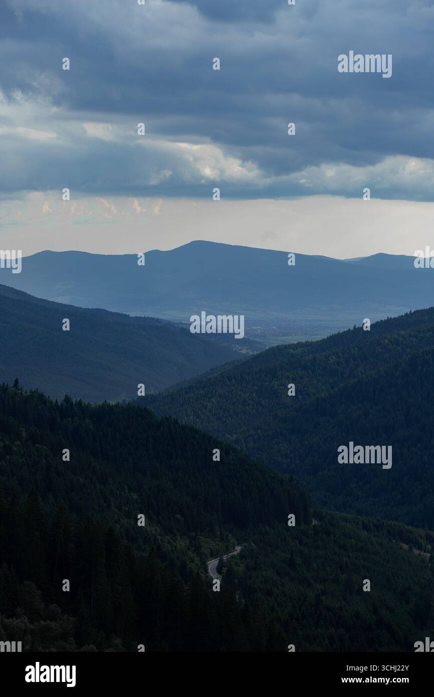 Les crêtes montagneuses ondulantes couvertes d'une forêt dense mènent l'œil vers une vallée et une ville lointaines, tandis qu'un ciel orageux dramatique se profile au-dessus de vous Banque D'Images