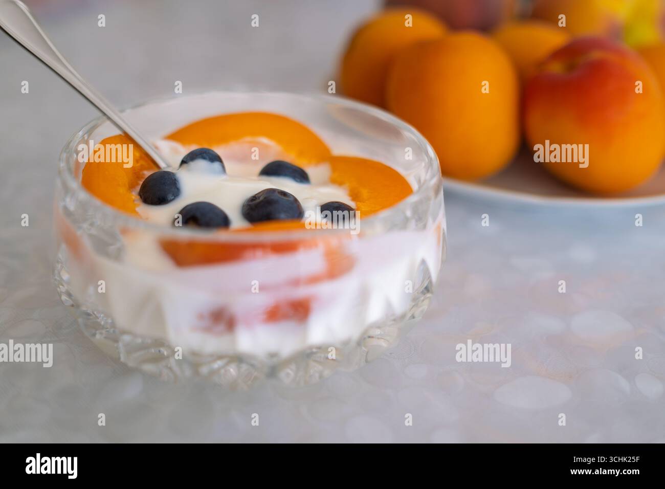 Bol en cristal rempli de yaourt crémeux, tranches d'abricots frais, et une pincée de bleuets se trouve sur une table, offrant un petit déjeuner nutritif. Banque D'Images