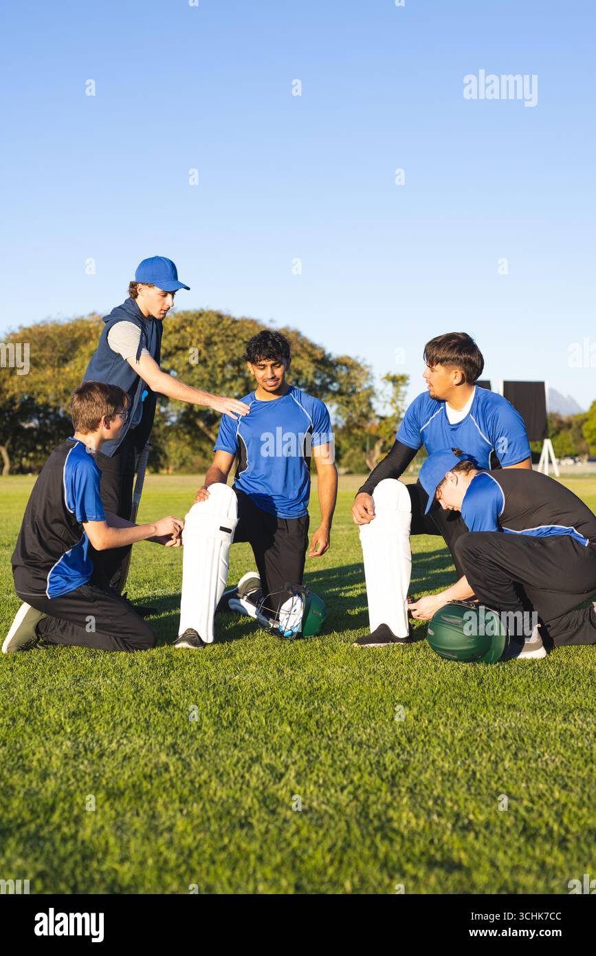 Équipe masculine diversifiée de cricket en maillots bleus attachant des coussinets de jambe par des souches, des casques sur le terrain de cricket Banque D'Images