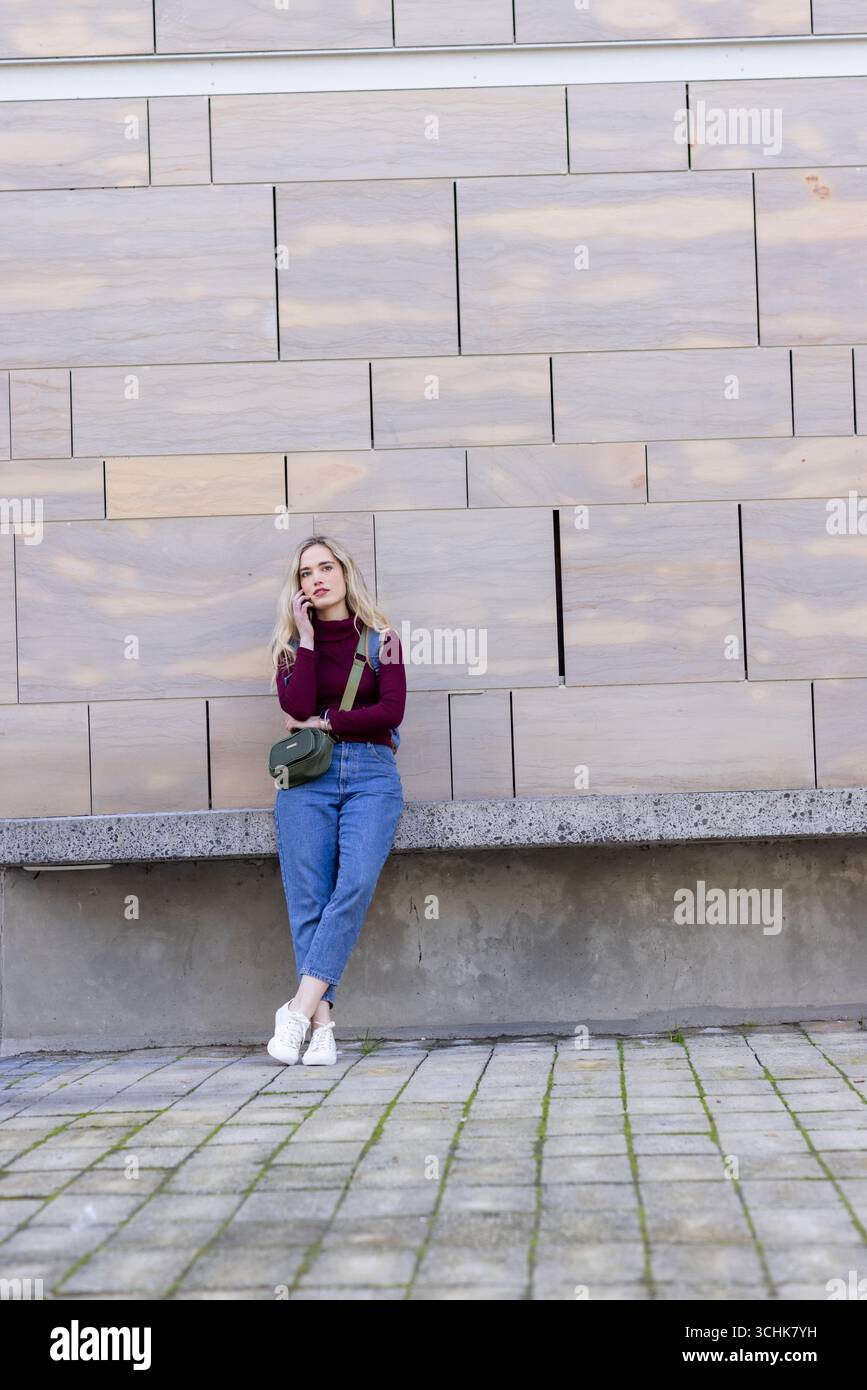 Femme appuyée sur le mur de panneau dans la place urbaine parlant sur smartphone, portant le sac vert olive Banque D'Images