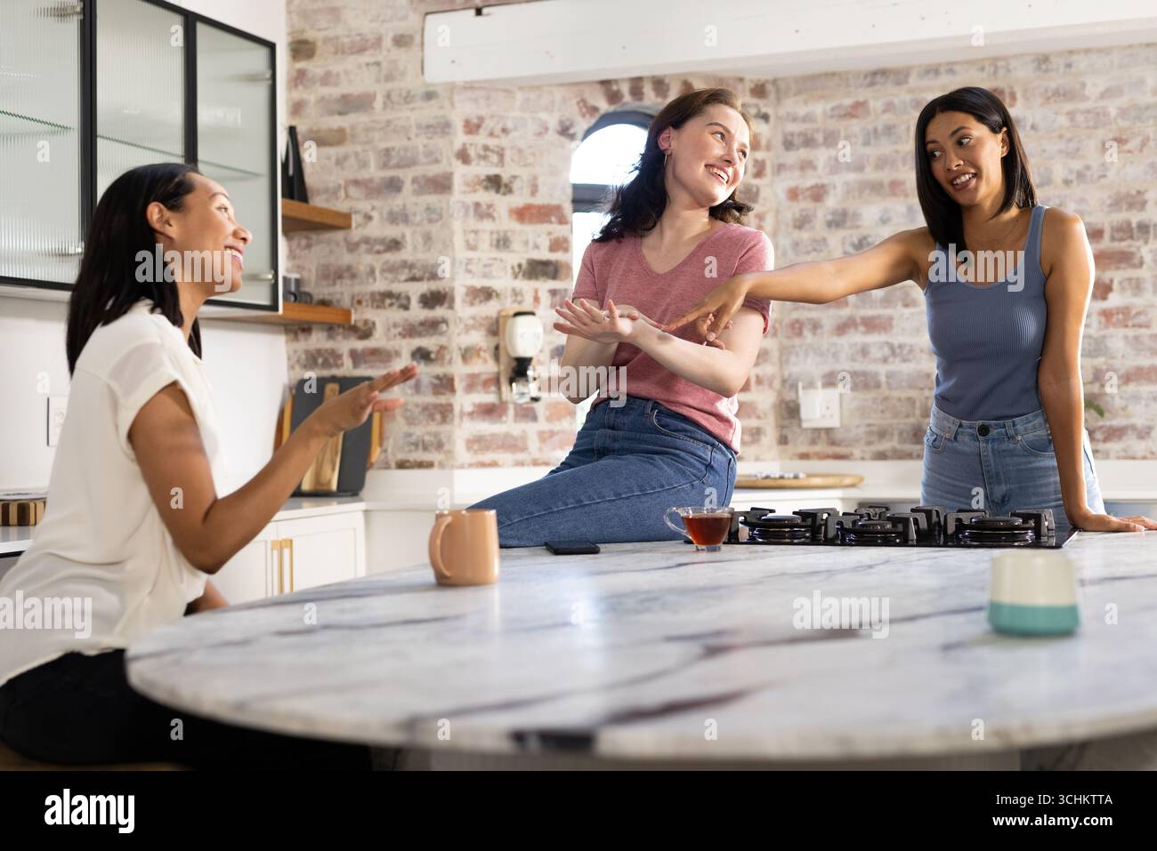 Riant et bavardant, trois jeunes femmes se rassemblent autour de l’îlot de cuisine Banque D'Images
