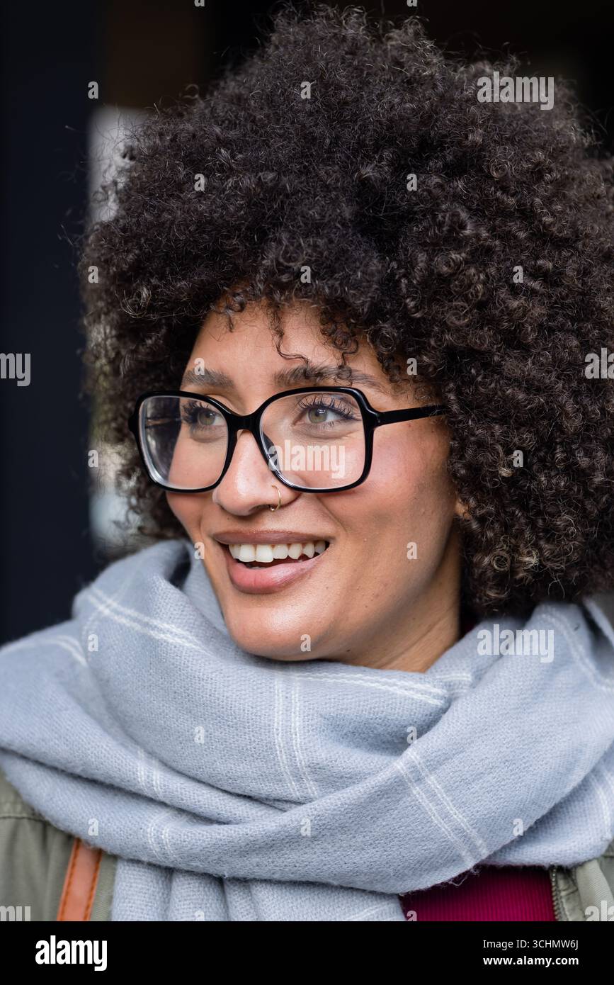 Femme portant des lunettes à cadre noir, anneau de nez, foulard gris souriant, regardant à gauche près de l'entrée Banque D'Images