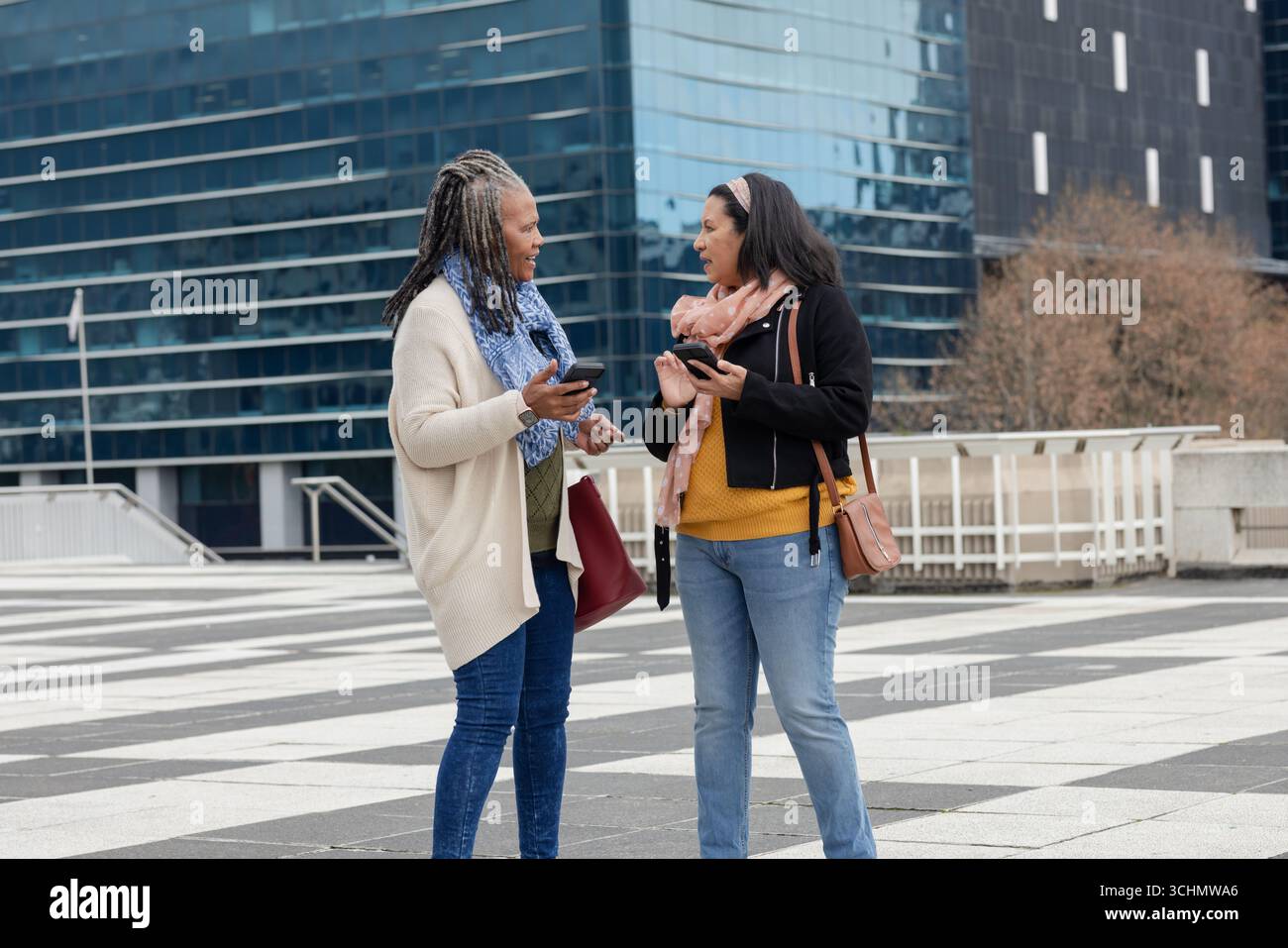 Diverses amies féminines se réunissant sur la plaza avant le bâtiment en verre tenant des smartphones, des sacs de transport Banque D'Images