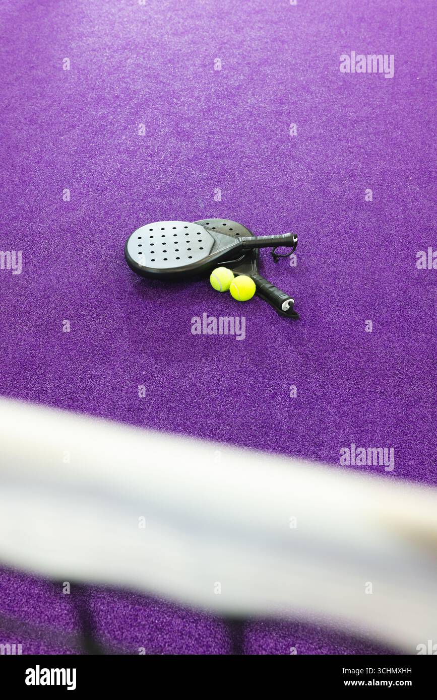 Deux raquettes de padel noir reposent sur un terrain en gazon violet à côté de boules jaunes à côté du filet flou Banque D'Images