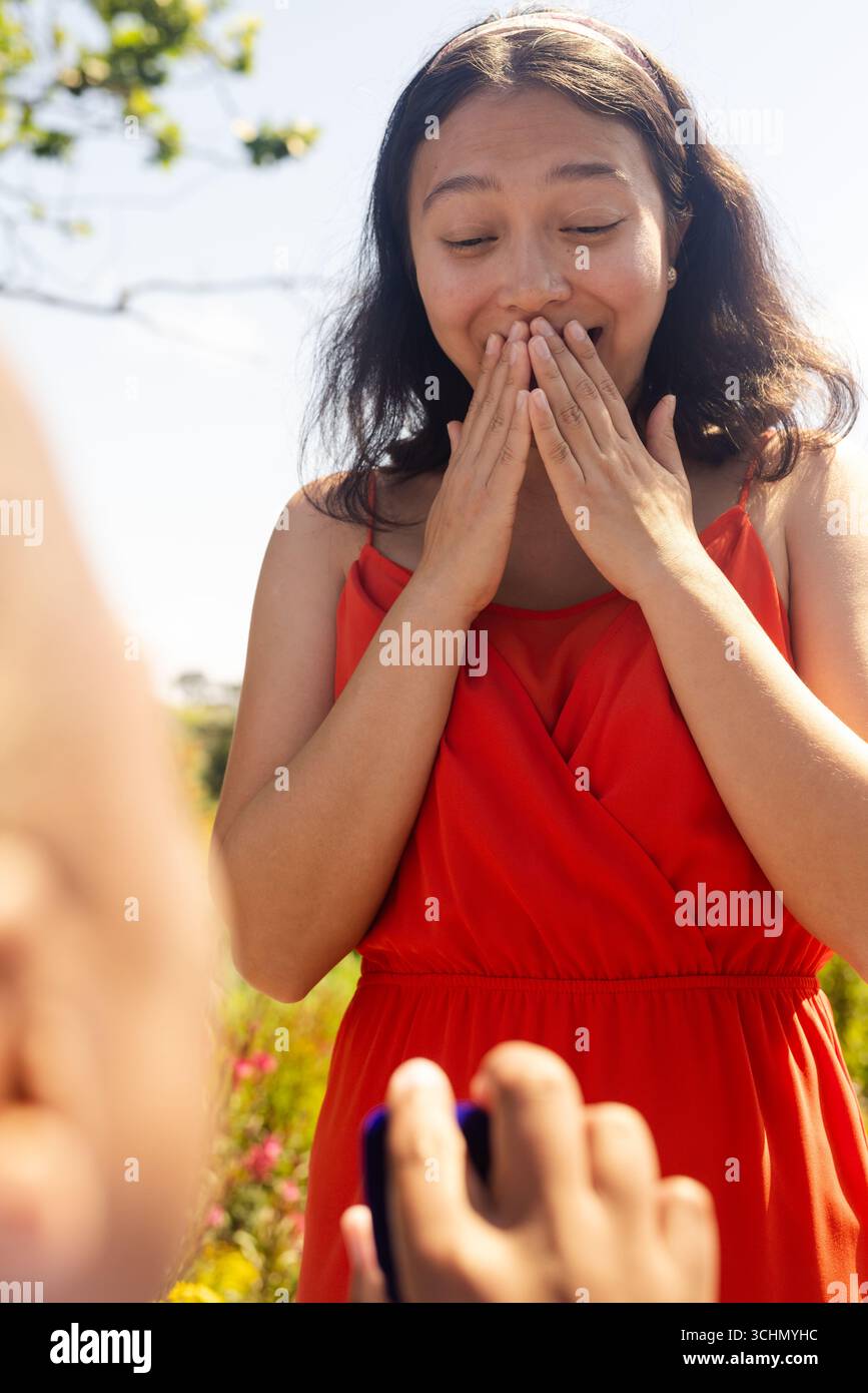 Femme surprise en robe rouge couvrant la bouche, recevant la proposition dans le jardin Banque D'Images