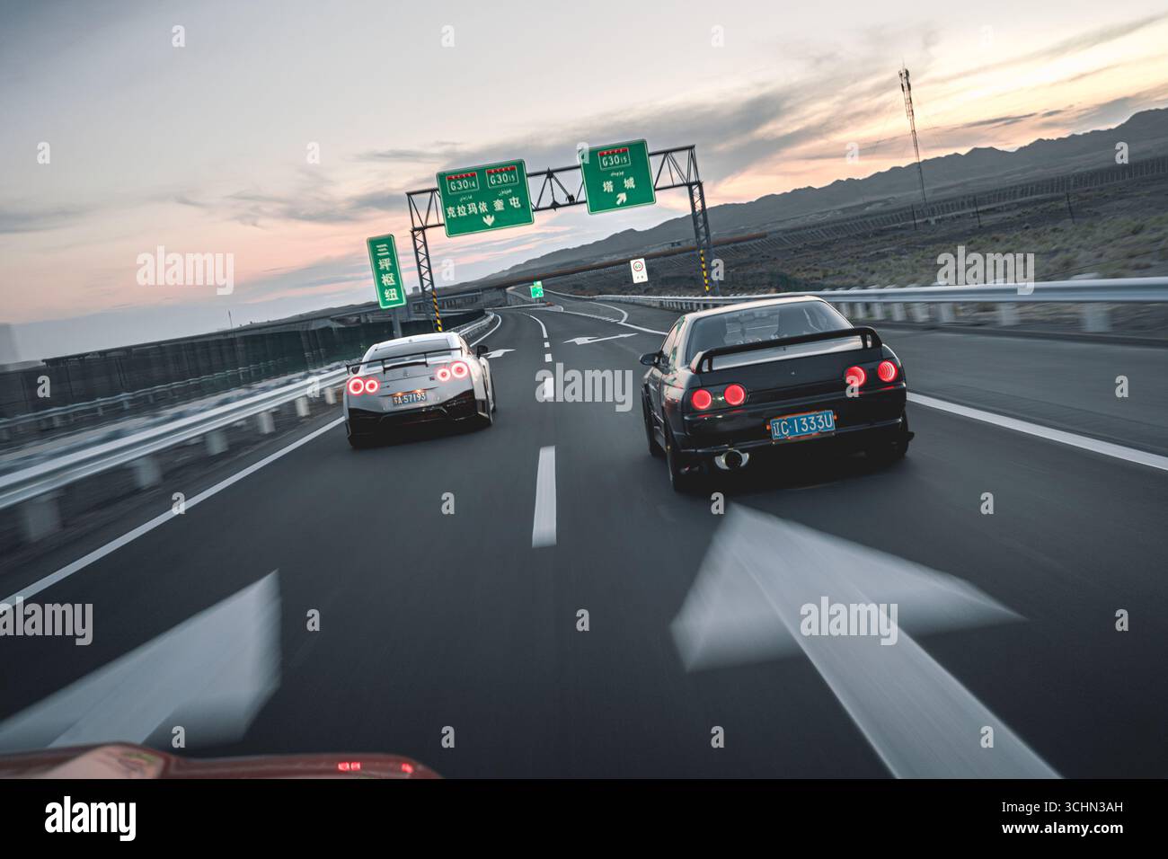 Une Nissan GT-R blanche et une Nissan Skyline noire conduisant côte à côte sur une autoroute à plusieurs voies sous un ciel de coucher de soleil. Banque D'Images