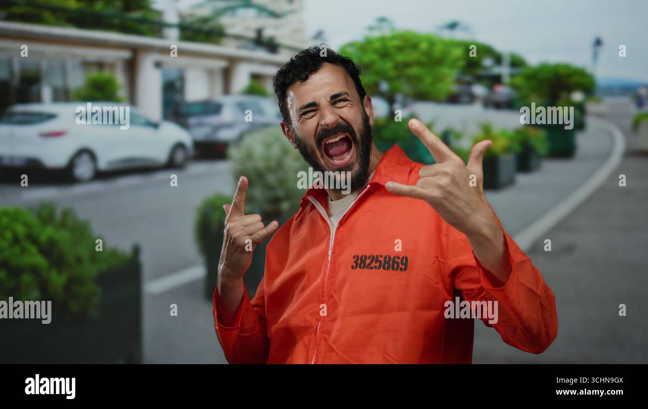 Homme hispanique avec une barbe dans une combinaison de prison orange fait un geste rock à l'extérieur dans une rue, mélangeant humour et rébellion dans un cadre vibrant CA Banque D'Images
