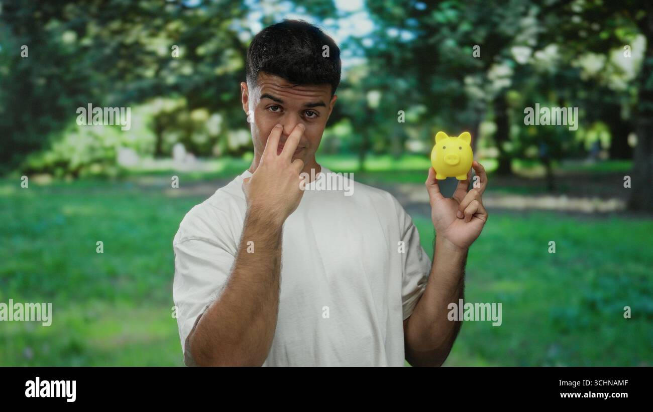 Jeune homme hispanique fait un geste tout en tenant une tirelire jaune dans un cadre de parc extérieur, portant une chemise blanche, transportant un joueur et observ Banque D'Images