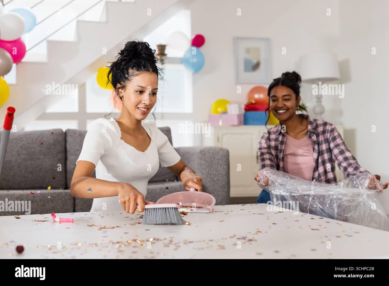Nettoyage après la fête, deux amies diverses rangeant le salon avec des sourires, à la maison Banque D'Images