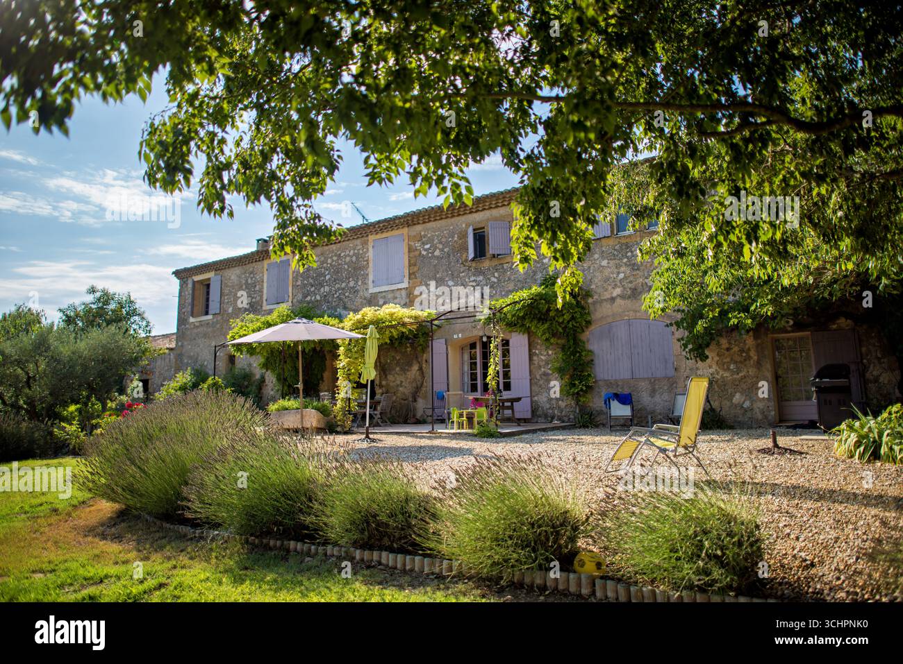Villa Patio Saint Martin de Crau France // SAINT-MARTIN-DE-Crau, France — Un patio en gravier avec mobilier de jardin et grand parasol est situé en face d'une villa du XVIIe siècle à Provence les Alpilles. Le bâtiment en pierre présente des volets violets sur ses fenêtres et ses portes. Une végétation luxuriante, comprenant des buissons de lavande et des arbres, entoure la propriété, avec une pelouse au premier plan. L'architecture de la villa reflète le style traditionnel de la région provençale. Banque D'Images