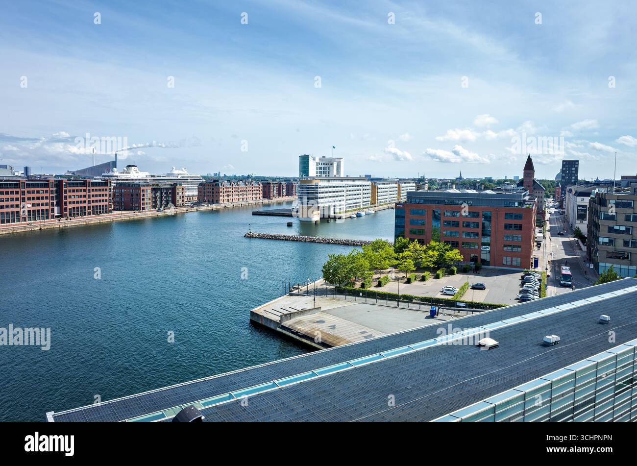 Architecture moderne et voie navigable à Christianshavn Copenhague Danemark // CHRISTIANSHAVN, Danemark — Un ferry DFDS est amarré dans le port de Christianshavn, un quartier de Copenhague, Danemark. La voie navigable est bordée d'une architecture moderne, y compris des bâtiments en briques et des structures en verre contemporaines. En arrière-plan, des éléments industriels tels que les cheminées sont visibles, à côté de la piste de ski distinctive de Copenhague et de la centrale électrique. La scène capture le mélange d'éléments maritimes historiques et de développement urbain moderne caractéristique de la capitale danoise. Banque D'Images