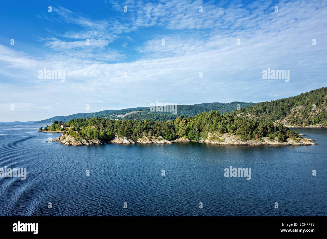Oslofjord Island with Trees Drøbak Sound Norway // OSLOFJORD, Norway — une île avec des arbres et des rives rocheuses est visible dans le Drøbak Sound, une partie de l'Oslofjord. Le fjord est une voie navigable majeure dans le sud-est de la Norvège, s'étendant à l'intérieur des terres à partir du détroit de Skagerrak. Le paysage présente des collines couvertes de forêts denses à feuilles persistantes, typiques de la côte norvégienne. L'eau affiche de légères ondulations, indiquant des conditions calmes. Banque D'Images