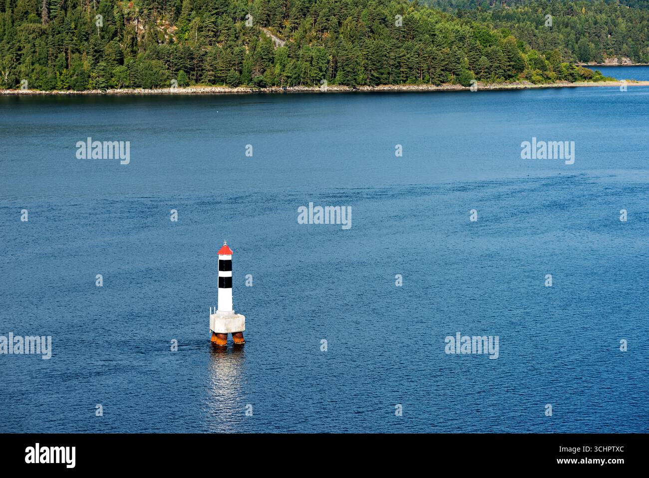 Bouée de navigation à Oslofjord Norvège // OSLOFJORD, Norvège — Une bouée de navigation se dresse dans les eaux de l'Oslofjord, un bras de mer profond qui s'étend de la côte norvégienne du Skagerrak au cœur de la région de la capitale du pays. La bouée, caractérisée par ses marques blanches, noires et rouges, joue un rôle crucial dans la sécurité maritime, guidant les navires à travers les voies navigables du fjord. Le fjord d'Oslofjord est une caractéristique géographique importante, connue pour sa beauté pittoresque, ses activités récréatives et son importance en tant que route maritime. La côte environnante est densément boisée, typique de la Norvège la Banque D'Images
