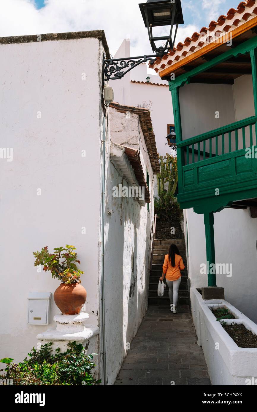 Une femme marche dans une ruelle étroite avec un balcon vert au-dessus d'elle. Le balcon est décoré d'une plante en pot et d'un vase. Banque D'Images