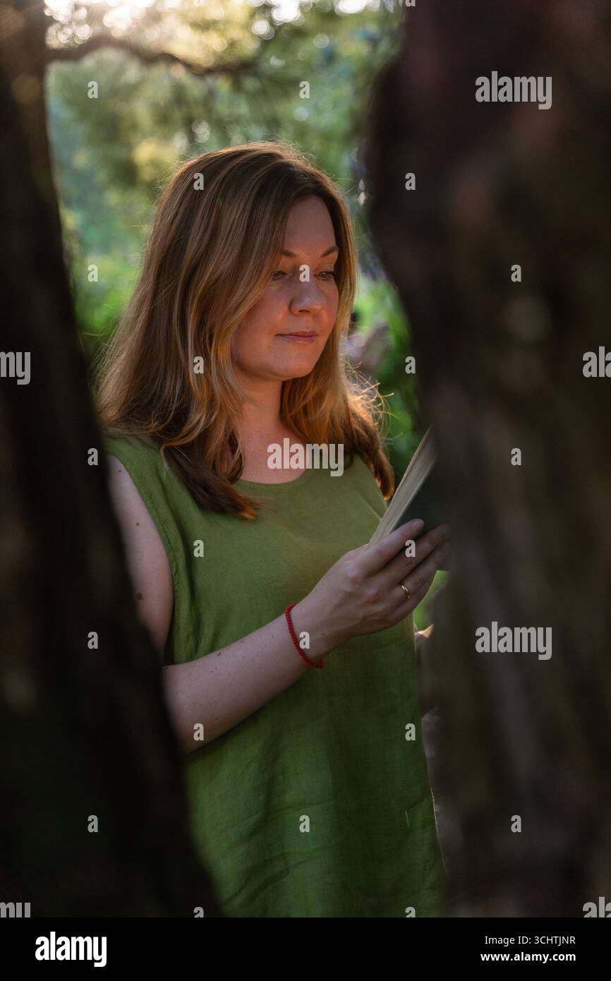 Une femme dans une blouse en lin vert lit un livre vintage à l'extérieur, encadré par des arbres dans une douce lumière du soir Banque D'Images