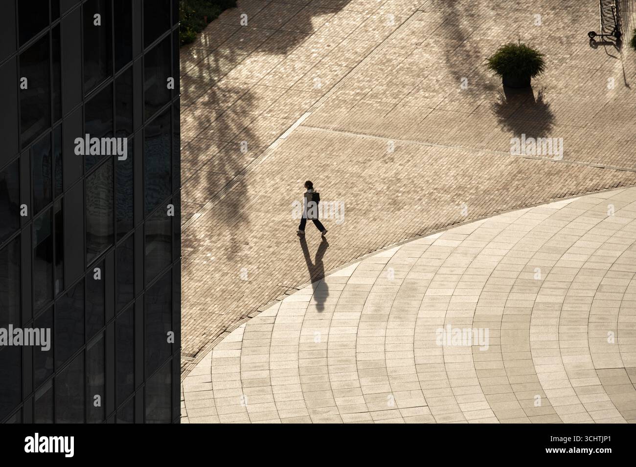 Une personne solitaire marche à travers une place urbaine ensoleillée, projetant une longue ombre sur le sol à motifs. La scène capture les lignes architecturales Banque D'Images
