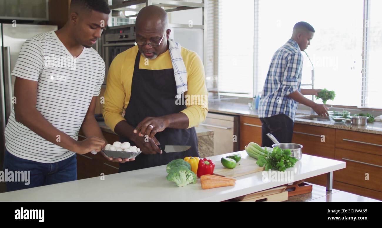 Tranchant le père dans la chemise jaune et le tablier, le fils coupant les champignons sur l'îlot de cuisine avec des légumes Banque D'Images