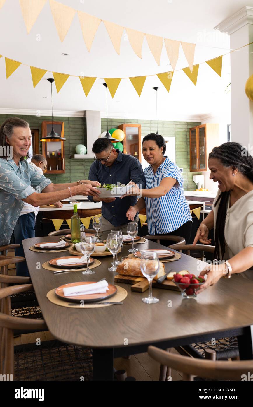 Divers amis seniors appréciant un repas de fête, mettant la table avec de la nourriture et des boissons à la maison Banque D'Images