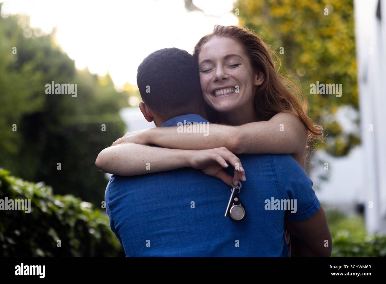 Jeune couple diversifié embrassant dans le jardin, femme souriant joyeusement tout en tenant les clés Banque D'Images