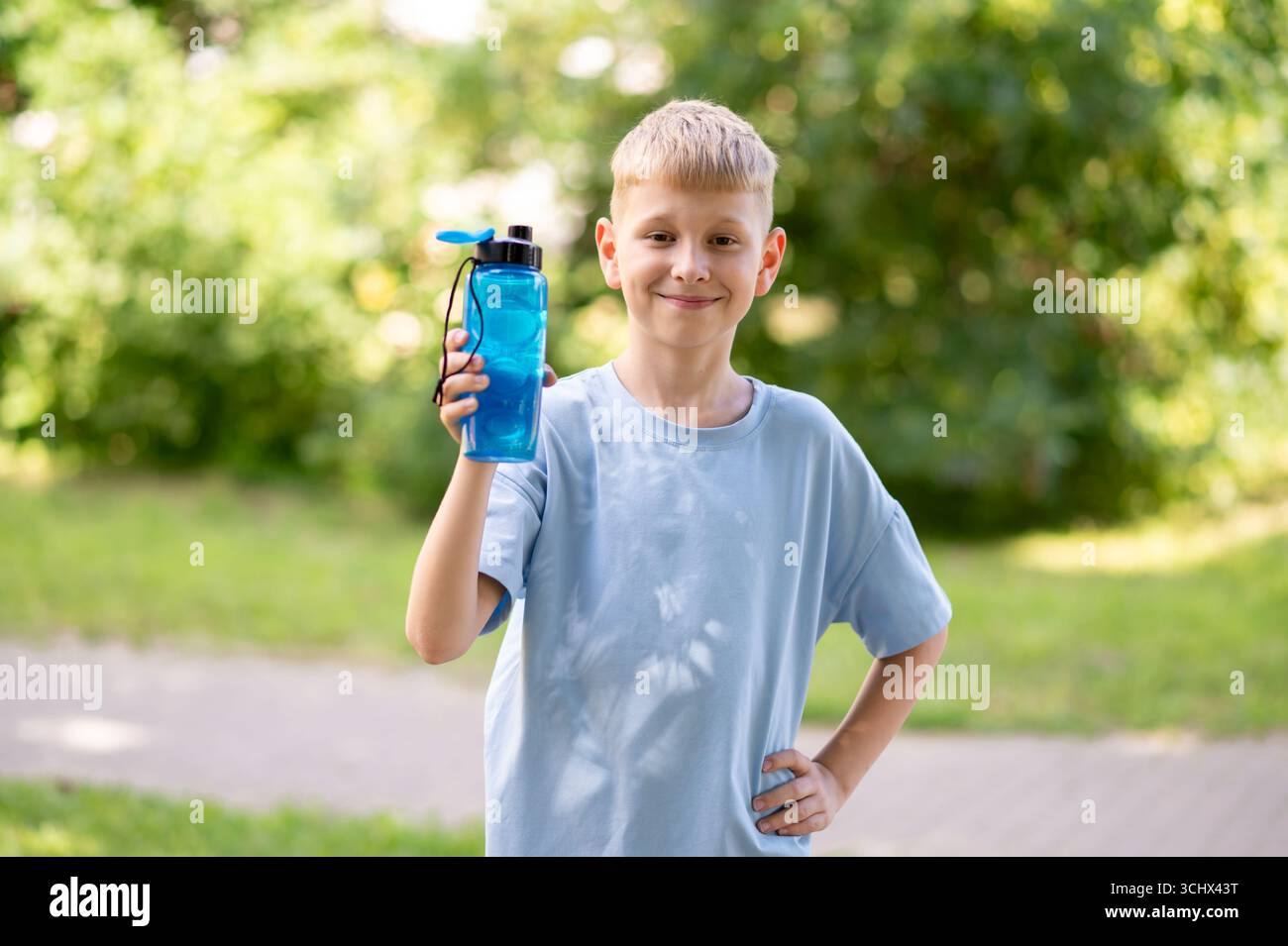 Garçon heureux tenant une bouteille d'eau tout en se tenant à l'extérieur, souriant et profitant d'une chaude journée ensoleillée avec de l'air frais et de l'énergie positive Banque D'Images