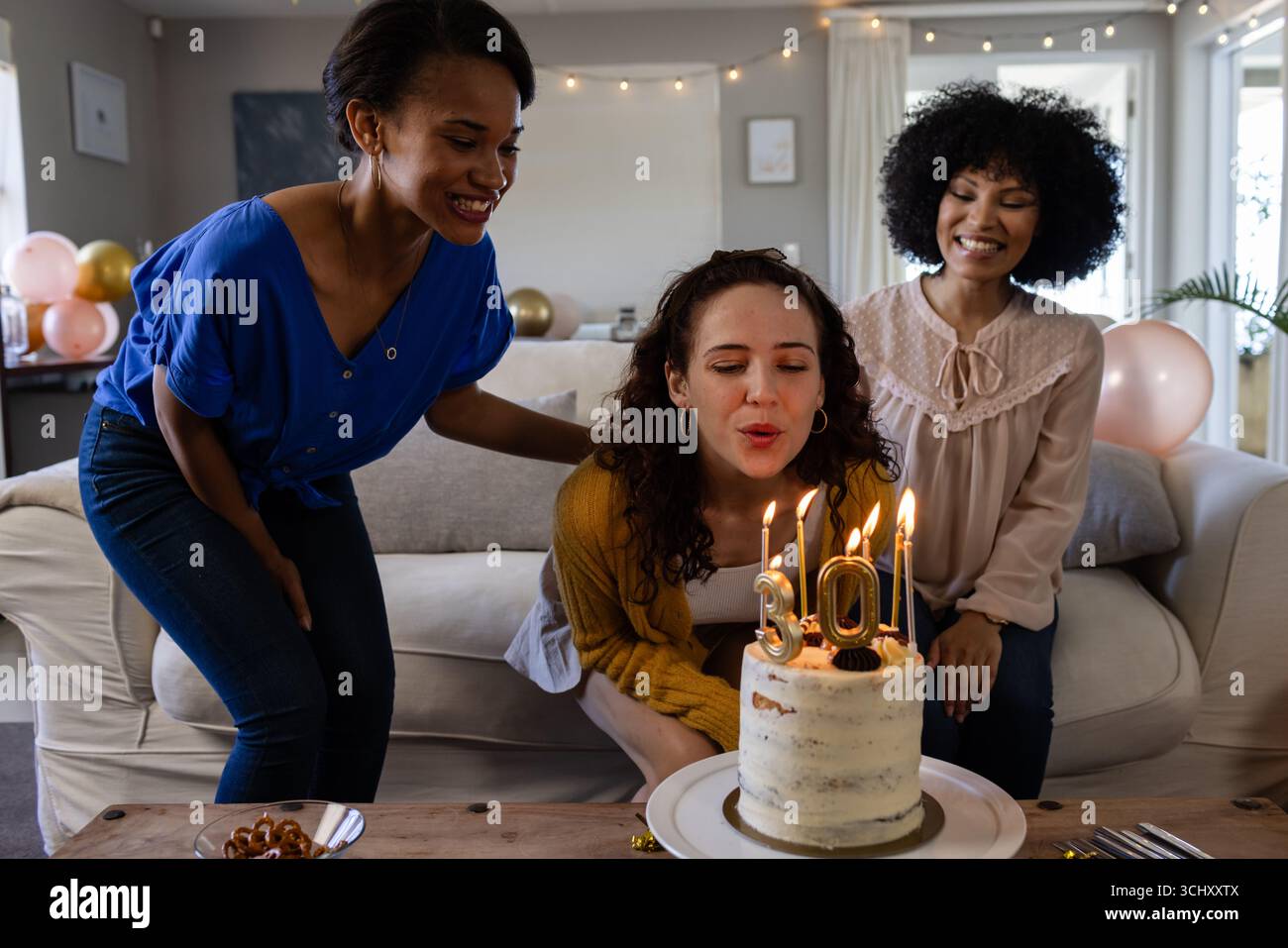 Diverses amies féminines célébrant l'anniversaire à la maison, femme soufflant des bougies sur le gâteau Banque D'Images