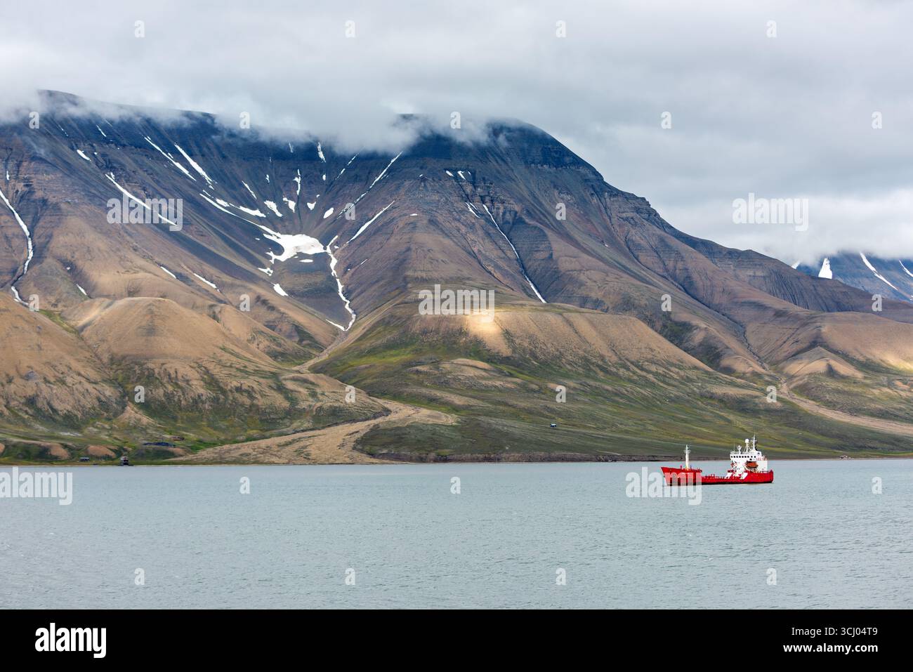 Red Cargo Ship voguant près des montagnes Longyearbyen Svalbard Norvège // LONGYEARBYEN, Svalbard — Un cargo rouge vole dans les eaux calmes du fjord, avec les montagnes escarpées et enneigées du Svalbard qui s'élèvent abruptement en arrière-plan. Le navire, une vue commune dans les eaux arctiques, navigue le long de la côte près de Longyearbyen, le plus grand village et centre administratif de l'archipel du Svalbard. Le paysage spectaculaire, caractérisé par des pentes arides et des parcelles de végétation verte, est typique de l'environnement arctique. Svalbard est connu pour son écosystème unique et est une destination populaire fo Banque D'Images