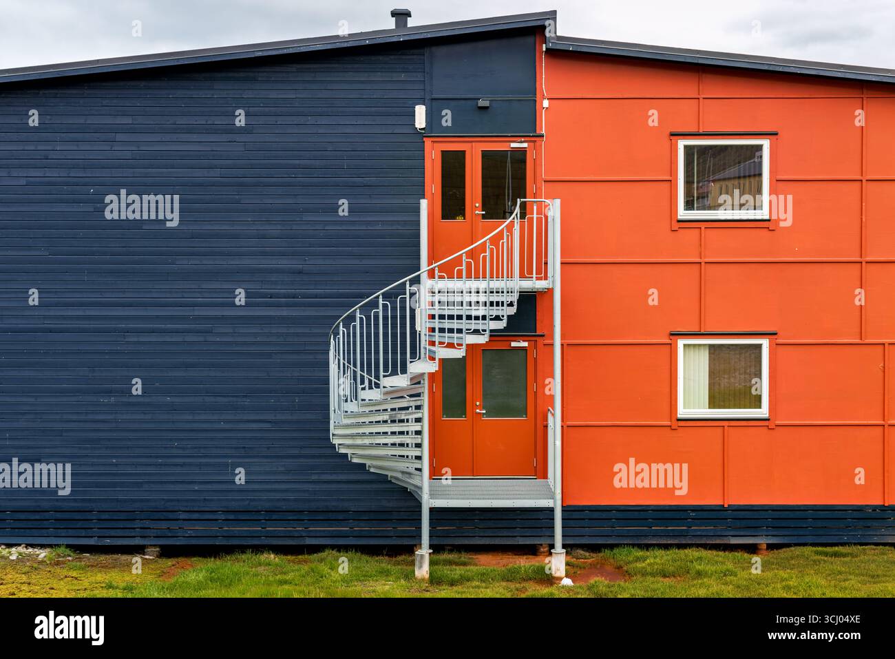 Bâtiment moderne avec escalier en spirale Longyearbyen Svalbard Norvège // LONGYEARBYEN, Svalbard — Un bâtiment moderne présente un contraste saisissant entre sa section bleu foncé, lambrissée horizontalement et sa façade orange vif, ponctuée de deux fenêtres. Un escalier en colimaçon extérieur argenté donne accès à des portes doubles sur le côté orange de la structure. Longyearbyen est le centre administratif et le plus grand village de l'archipel du Svalbard, situé dans l'océan Arctique. L'archipel est un territoire norvégien situé entre la Norvège continentale et le pôle Nord. Banque D'Images