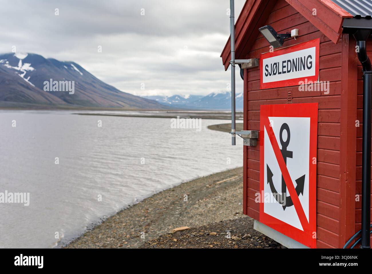 Sjoledning panneau Longyearbyen Svalbard Norvège // LONGYEARBYEN, Svalbard — Une structure en bois rouge avec un panneau indiquant « SJØLEDNING » est visible à Longyearbyen, Svalbard. Le bâtiment présente également un panneau rouge et blanc avec un symbole d'ancre noire barrée, indiquant une interdiction. En arrière-plan, un plan d'eau calme s'étend vers des montagnes enneigées sous un ciel couvert. Longyearbyen est la plus grande colonie et centre administratif de l'archipel du Svalbard, situé dans l'extrême-Arctique. Le terme « Sjøledning » se traduit par « leadership maritime » ou « leadership maritime » en norvégien, su Banque D'Images