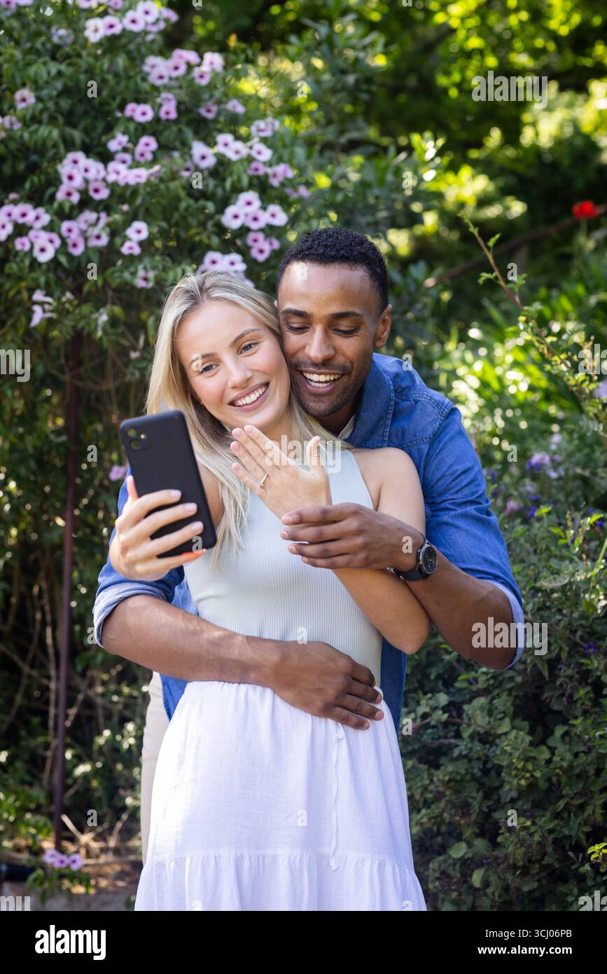 Couple diversifié célébrant l'engagement en prenant selfie dans le jardin, montrant le bonheur et l'amour Banque D'Images