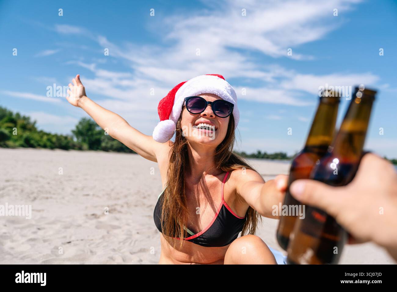 Femme joyeuse dans le chapeau de Santa acclamant avec bouteille de bière sur la plage tropicale Banque D'Images
