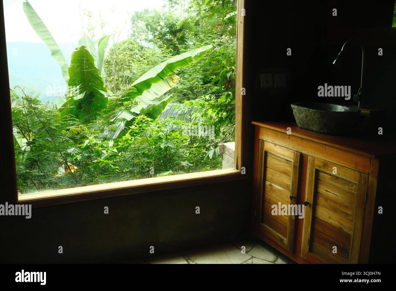 Une salle de bain rustique avec un évier en pierre naturelle, armoire en bois, avec une vue sur la Jungle à travers une large fenêtre Banque D'Images
