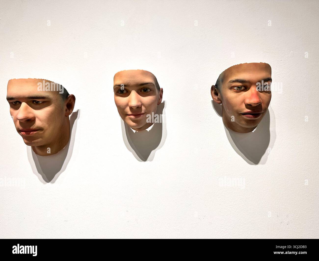 Trois visages imprimés en 3D réalistes montés sur un mur blanc, créant un affichage visuel saisissant avec des ombres. Banque D'Images