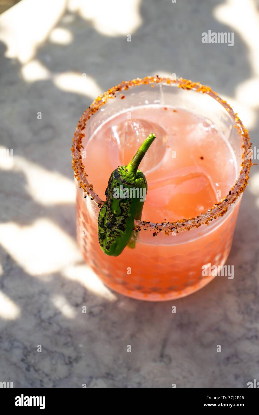 Cocktail rose rafraîchissant servi dans un verre texturé avec des glaçons, garni d'un piment vert carbonisé et d'un bord assaisonné de Chili-lime. Banque D'Images