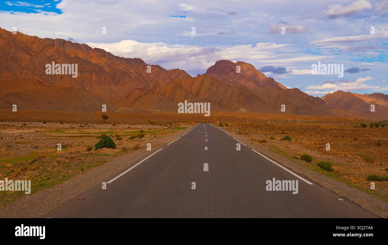 Autoroute vide au Maroc avec voiture lointaine conduisant vers des montagnes rocheuses spectaculaires et paysage désertique sous ciel nuageux du soir Banque D'Images