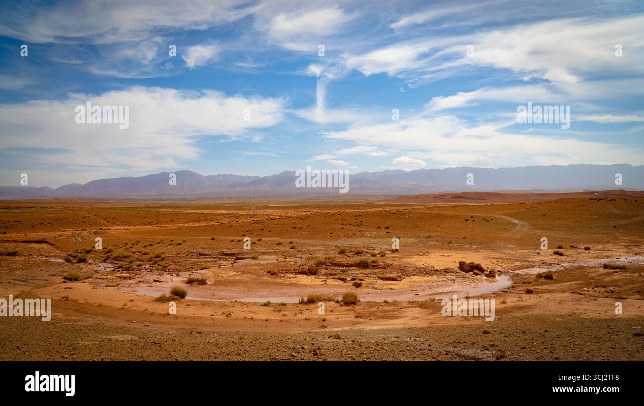 Vaste plaine désertique aride au Maroc avec des arbustes éparpillés, lit de rivière sec et montagnes lointaines de l'Atlas sous ciel bleu avec des nuages blancs dérivants Banque D'Images