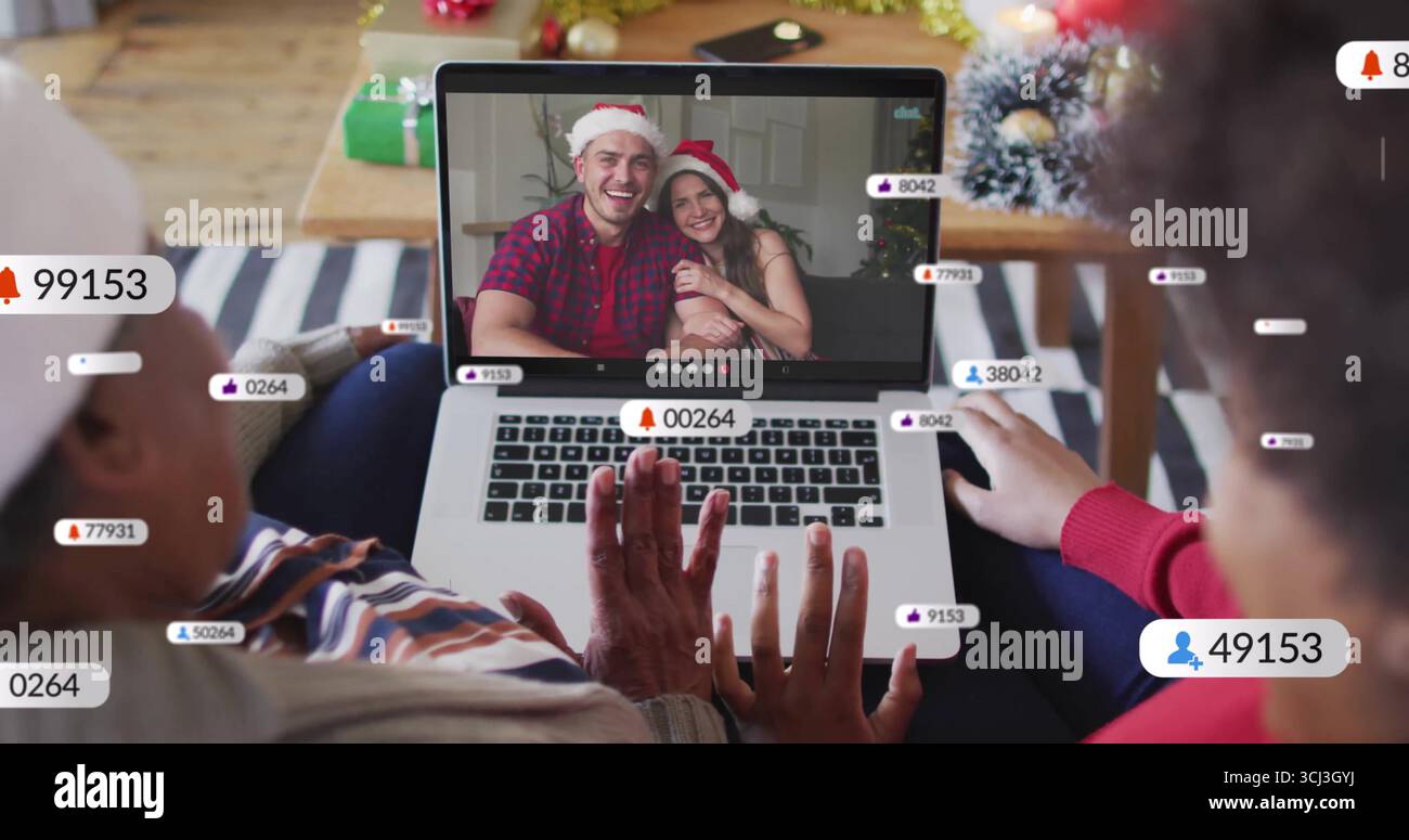 Agitant des femmes afro-américaines saluant le couple de chapeau de Père Noël à l'aide d'un ordinateur portable à la maison, avec des alertes de streaming. Festif, célébration, numérique, saisonnier, chaleur, Banque D'Images
