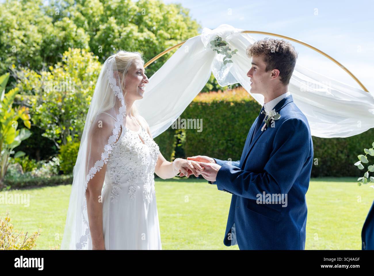 Mariée et mariée échangeant leurs vœux à l'extérieur sous une élégante arche florale, souriant chaleureusement Banque D'Images
