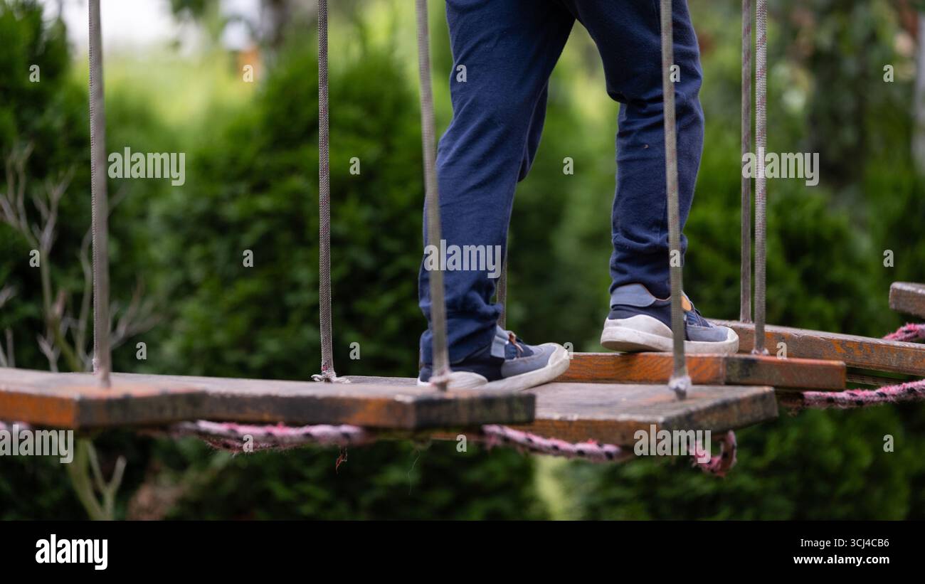 Gros plan des jambes d un enfant debout sur un pont de corde en bois dans un parc d aventure en plein air. L'image illustre l'équilibre, le mouvement et l'activité de l'enfance Banque D'Images
