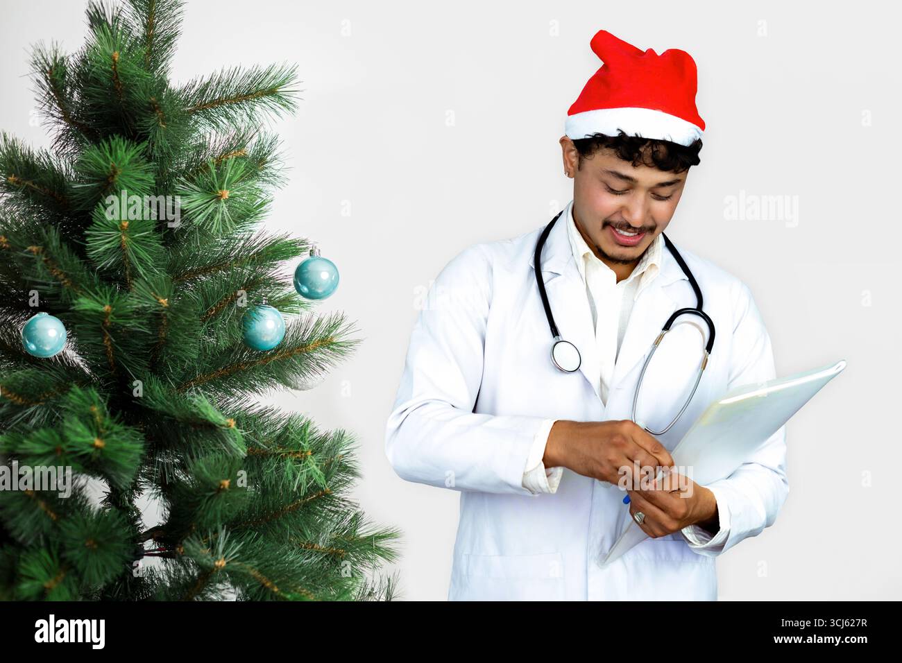 Beau médecin masculin portant un chapeau de Père Noël, debout devant un arbre de Noël avec des cadeaux, célébrant la saison des fêtes dans un setti de soins de santé Banque D'Images
