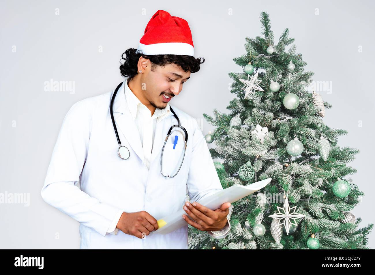 Beau médecin masculin portant un chapeau de Père Noël, debout devant un arbre de Noël avec des cadeaux, célébrant la saison des fêtes dans un setti de soins de santé Banque D'Images