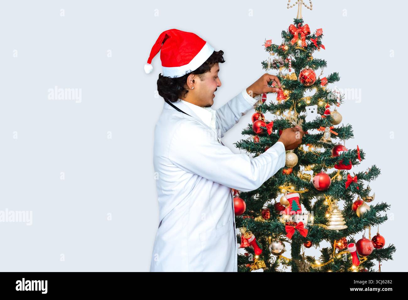 Beau médecin masculin portant un chapeau de Père Noël, debout devant un arbre de Noël avec des cadeaux, célébrant la saison des fêtes dans un setti de soins de santé Banque D'Images