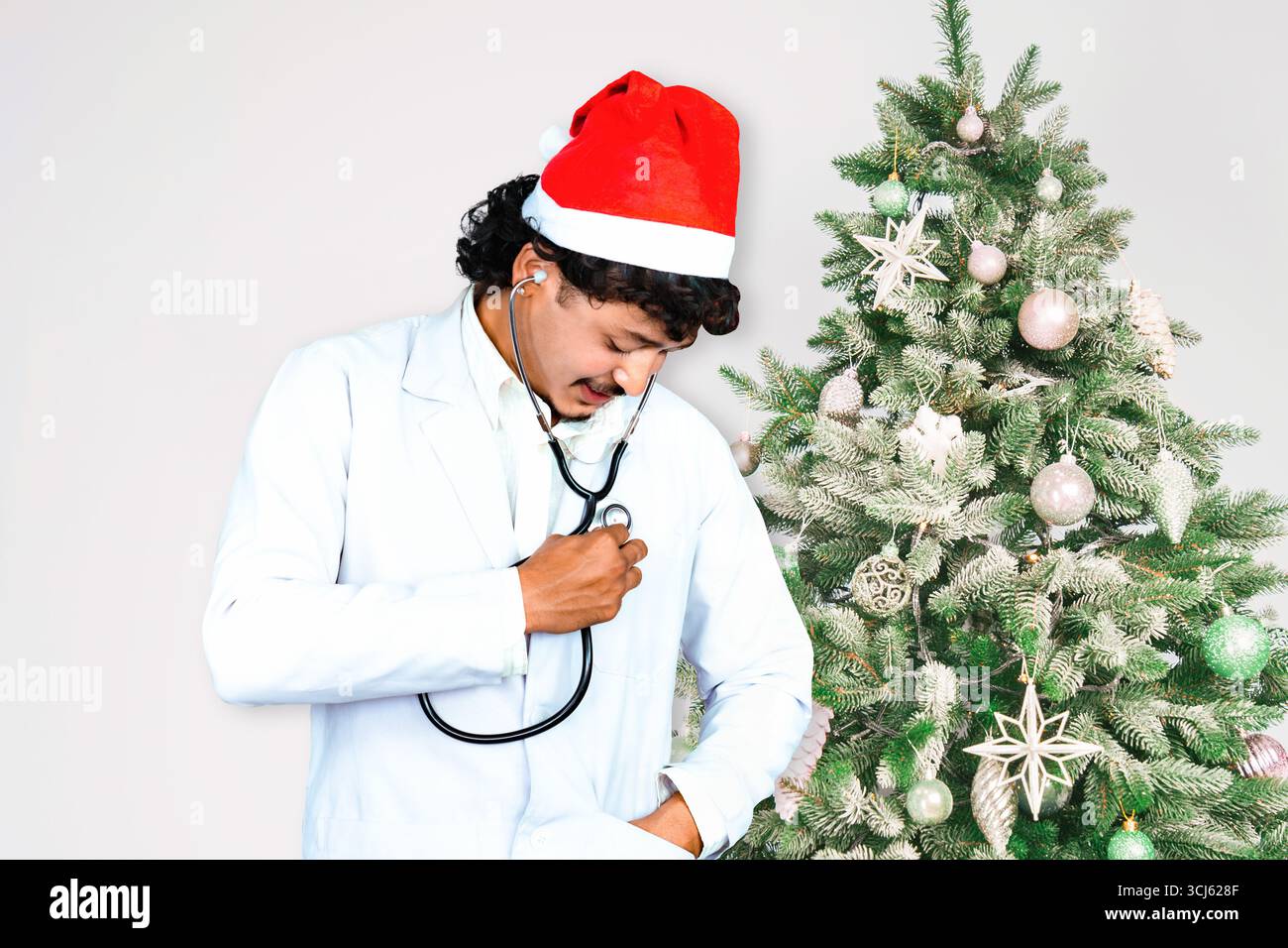 Beau médecin masculin portant un chapeau de Père Noël, debout devant un arbre de Noël avec des cadeaux, célébrant la saison des fêtes dans un setti de soins de santé Banque D'Images
