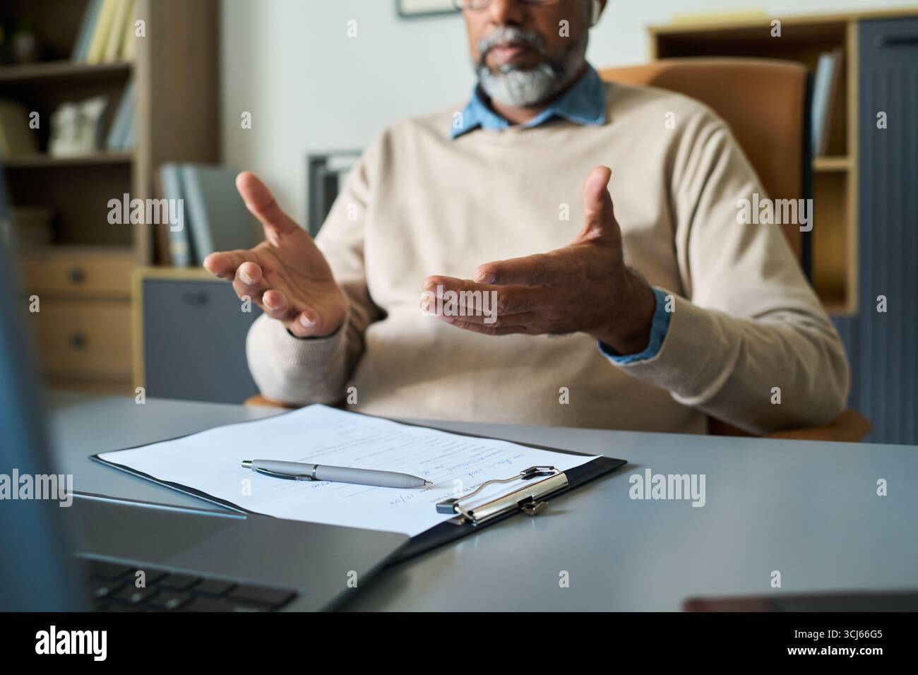 Homme indien d'âge moyen faisant des gestes avec les mains tout en étant assis à son bureau pendant une réunion d'affaires, document et stylo sur le presse-papiers au premier plan, visage partiellement visible, portant des lunettes Banque D'Images