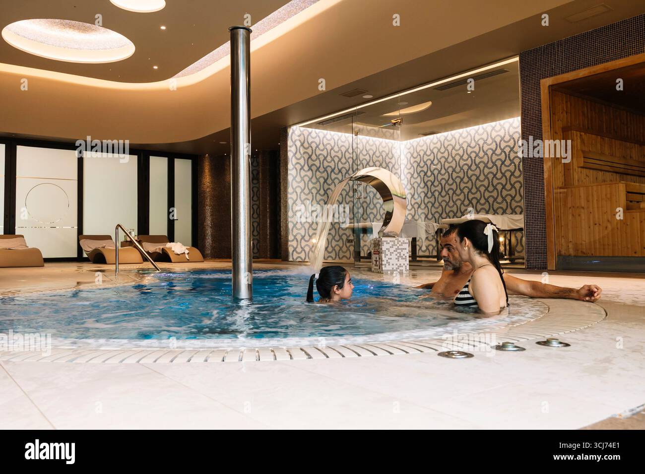 Une famille profite d'un moment de détente dans une piscine. La piscine est entourée d'un mur blanc et a une fontaine au milieu. La famille est assise dans le Banque D'Images