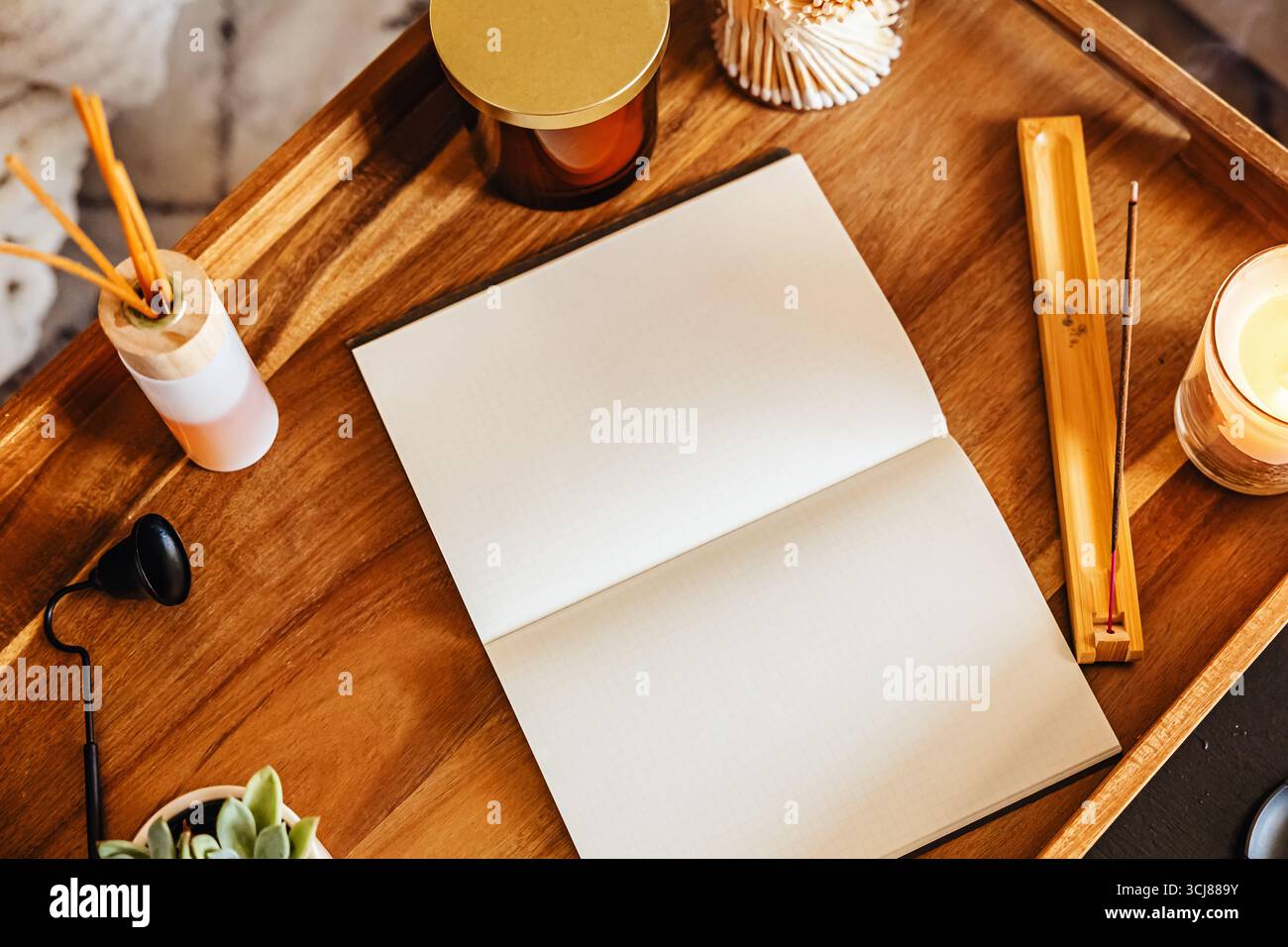 Espace de travail confortable avec cahier ouvert pour journaliser sur un plateau en bois entouré de bougies parfumées, encens et une petite plante. Banque D'Images