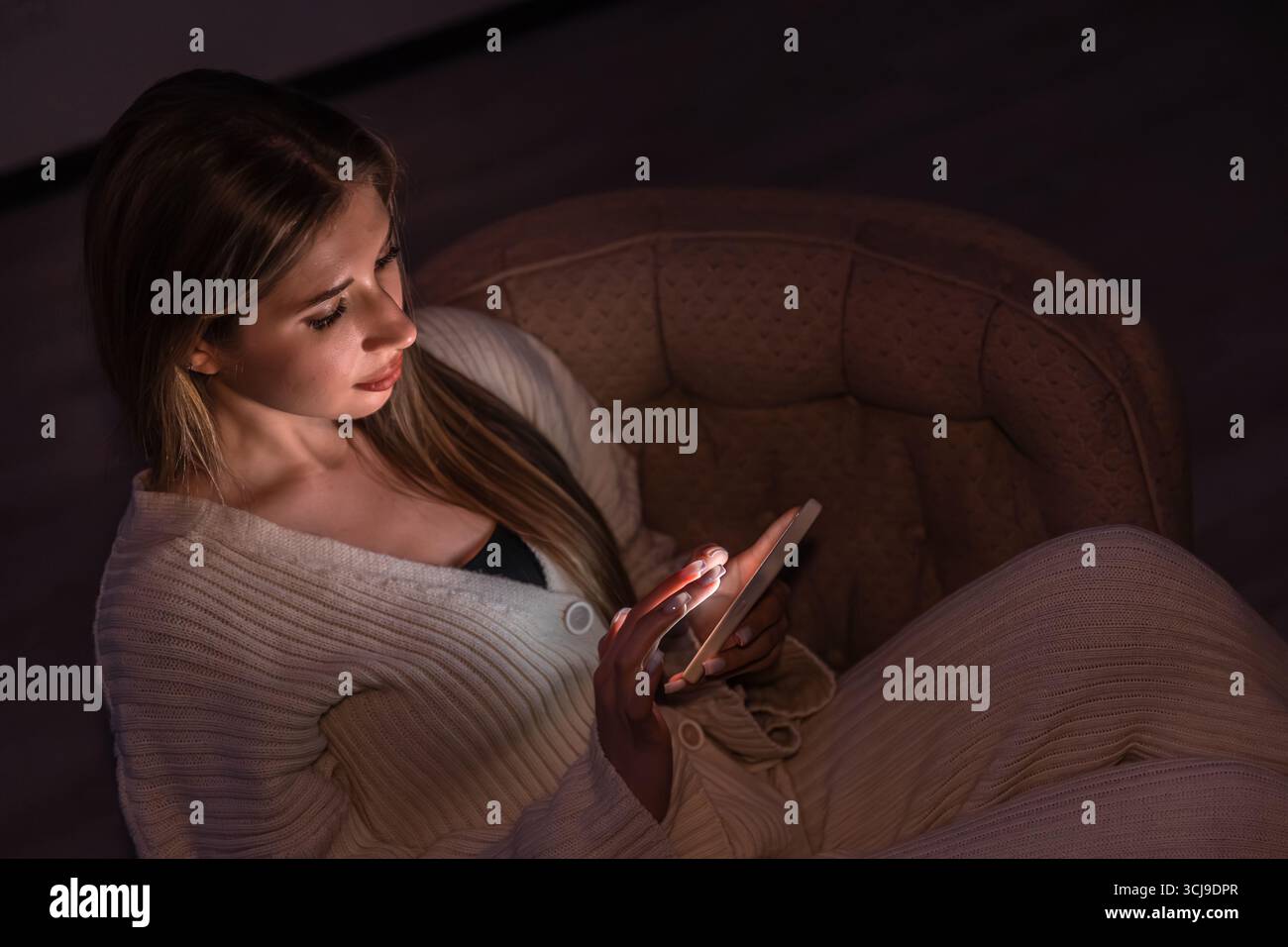 Jeune femme en fauteuil parcourant son téléphone par une lumière chaude. Ambiance chaleureuse pour la maison. Banque D'Images